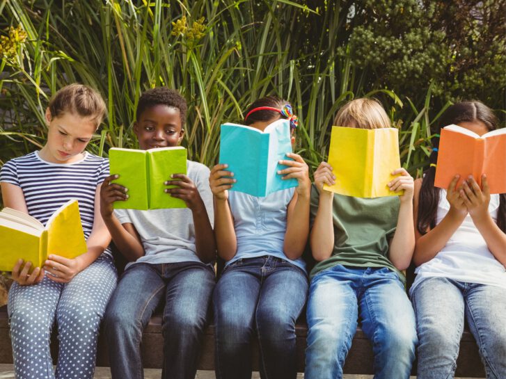 Fünf Kinder sitzen auf einer Bank und jede Person hat ein Buch in der Hand und liest