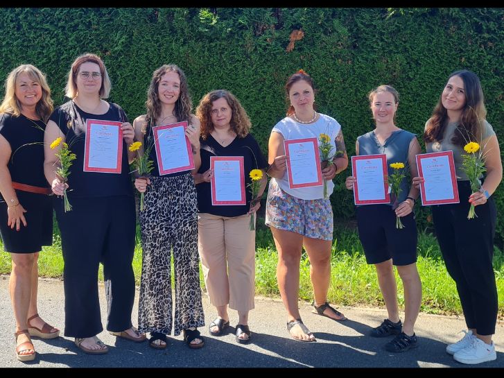 Sieben Frauen stehen nebeneinander im Freien vor einer Hecke und halten ihre Abschlusszertifikate hoch.