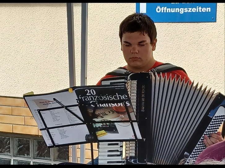 Ein Junge mit Akkordeon sitzt vor dem Gebäude der Gemeinwesenarbeit Bürstadt und spielt Französische Chansons.