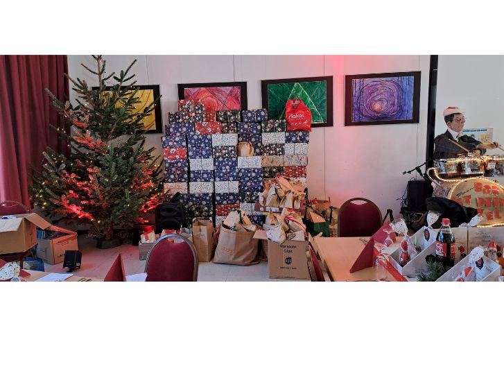 Im Gemeindehaus der St. Laurentius Kirche in Bensheim steht links ein noch ungeschmückter Weihnachtsbaum. Daneben stapeln sich Geschenkpakete und Geschenktüten. Links befindet sich ein Schlagzeug, an dem eine Puppe namens Onkel Bob sitzt. Im Hintergrund hängen vier bunte Bilder an der Wand. Im Vordergrund ist ein Teil der Tischdeko zu sehen.