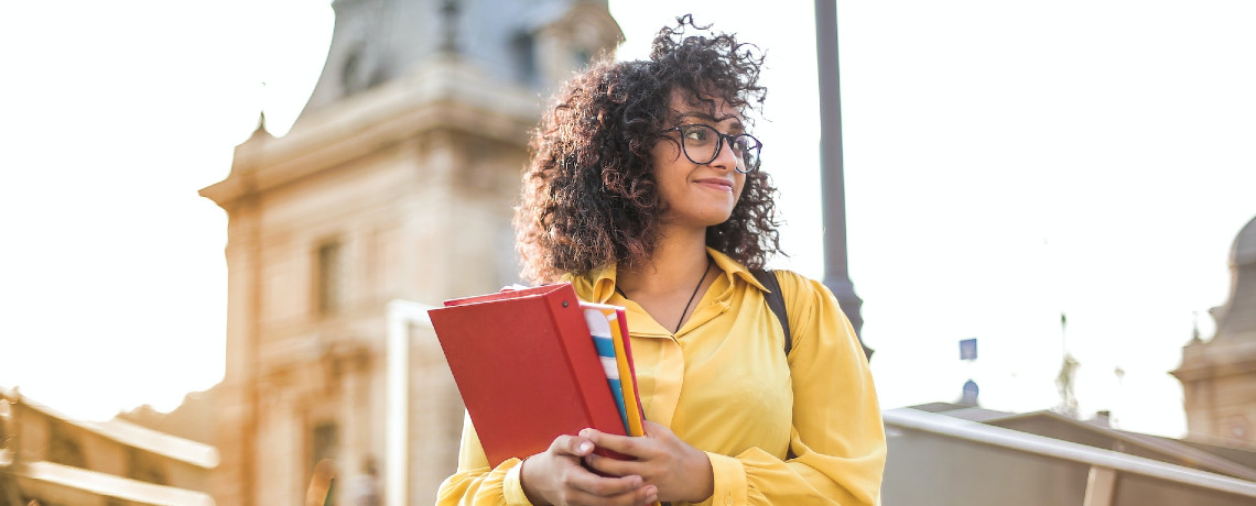 Auf dem Foto ist eine junge Frau mit gelber Bluse zu sehen. Sie trägt eine Brille und hält in den Händen eine Mappe und einen Stift. Im Hintergrund ist eine Kirche.