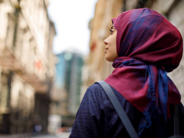 Junge Frau mit Kopftuch blickt in Richtung einer Häuserfront, die verschwommen zu sehen ist.